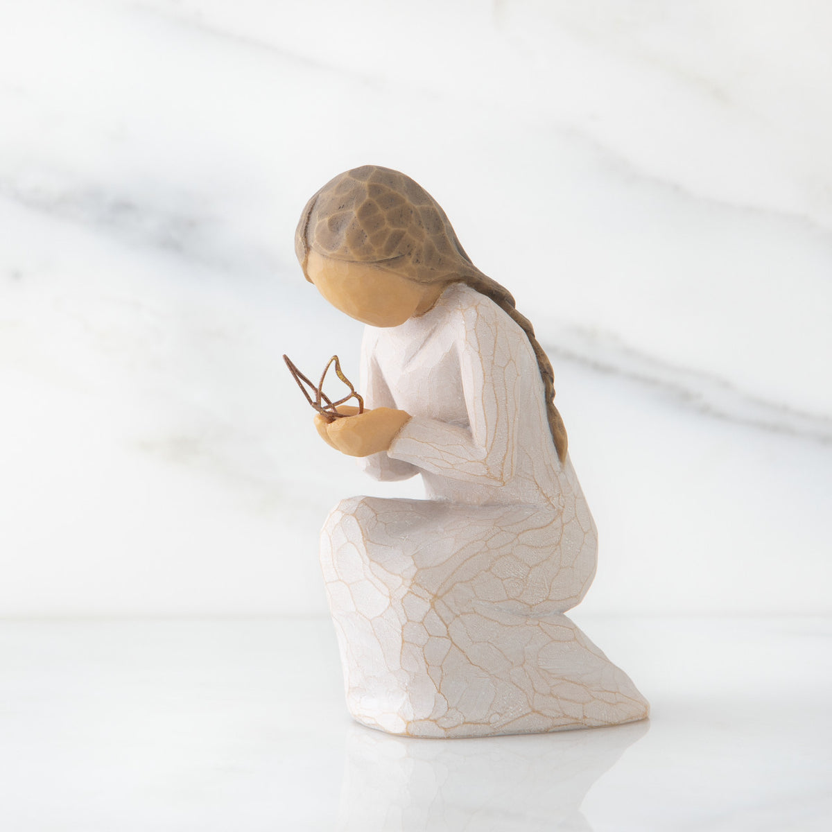Side view of kneeling female figure in cream dress, with wire butterfly on extended hands, on marble background
