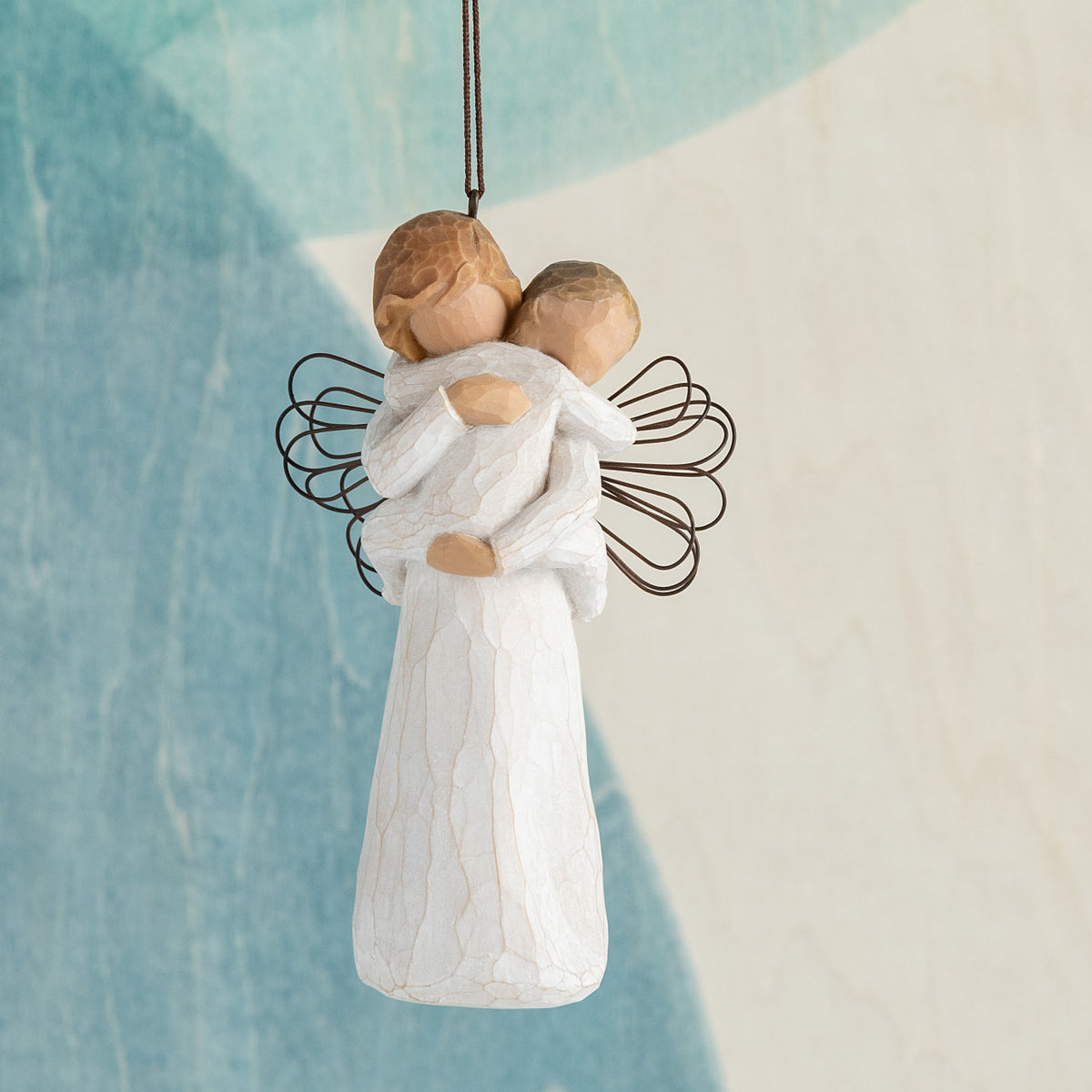 Willow Tree Angel's Embrace Ornament, 5"h hand-painted resin figure in cream dress with wire wings, holding baby in arms, brown elastic cord, hanging in front of blue painting