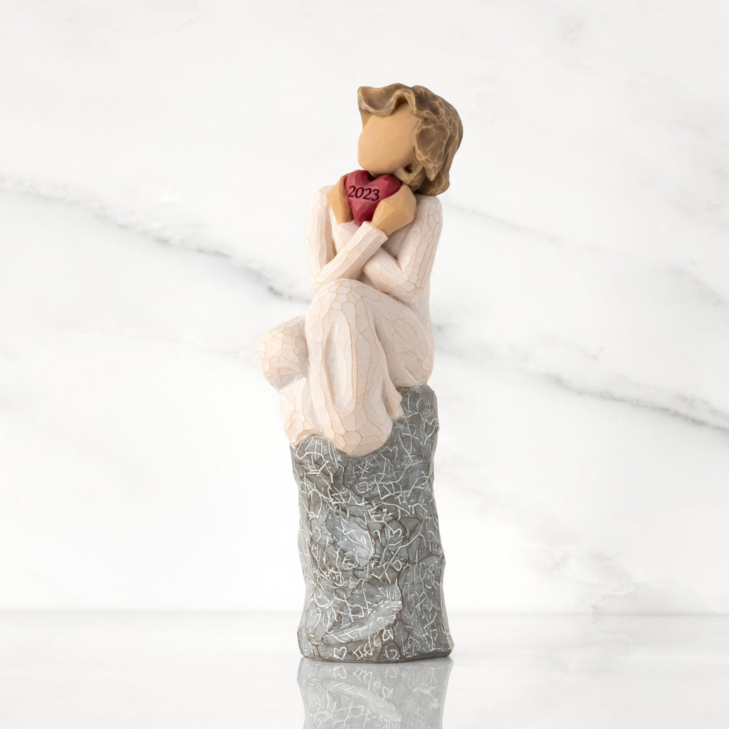 Front view: figure in cream dress, seated on gray rock, knees drawn up to chest, holding red heart engraved with year 2023
