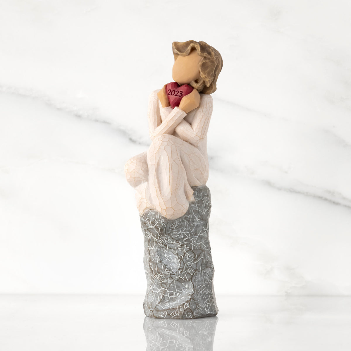 Front view: figure in cream dress, seated on gray rock, knees drawn up to chest, holding red heart engraved with year 2023