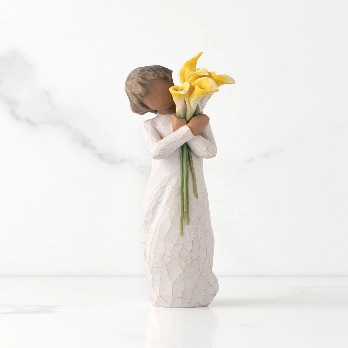 Front view of standing figure in cream dress with darker skin holding yellow calla lilies in her arms