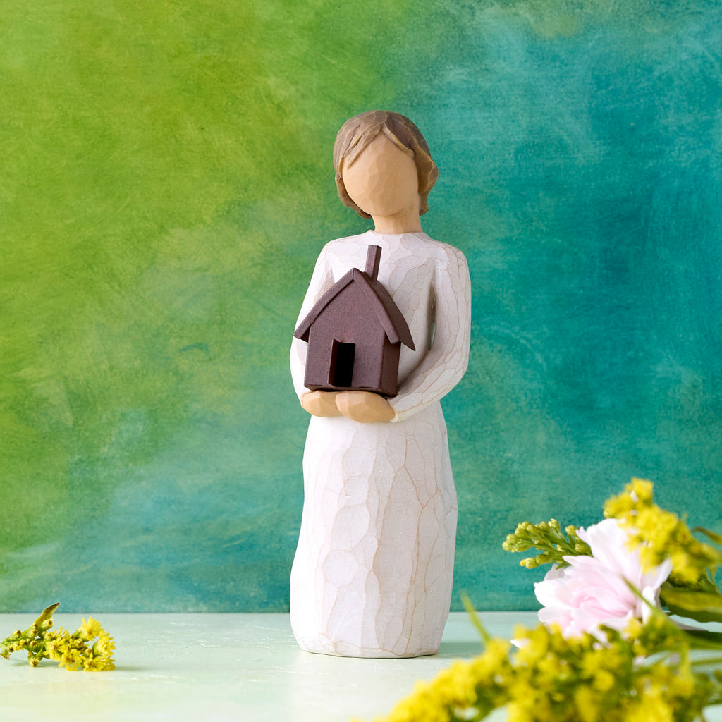 Willow Tree Mi Casa, 5.5”h hand-painted standing resin figure in cream dress, holding dimensional metal house in her arms, in front of deep teal painting