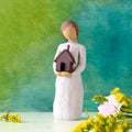Willow Tree Mi Casa, 5.5”h hand-painted standing resin figure in cream dress, holding dimensional metal house in her arms, in front of deep teal painting