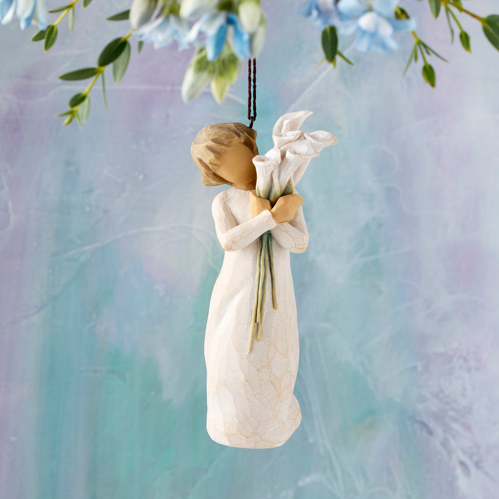 Willow Tree Beautiful Wishes Ornament, 4.5”h hand-painted resin figure in cream dress holding bouquet of white calla lilies, brown elastic cord, hanging in front of blue painting
