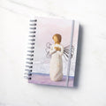 Front view: spiral-bound notebook featuring front of figure with wire wings in cream dress carved with natural symbols, hands crossed under gold heart on chest