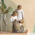 Willow Tree Generations, 7”h hand-painted resin figure of seated older woman sewing blue quilt on her lap; standing woman at her shoulder; seated young girl at her feet, all in neutral clothing, on table with green florals