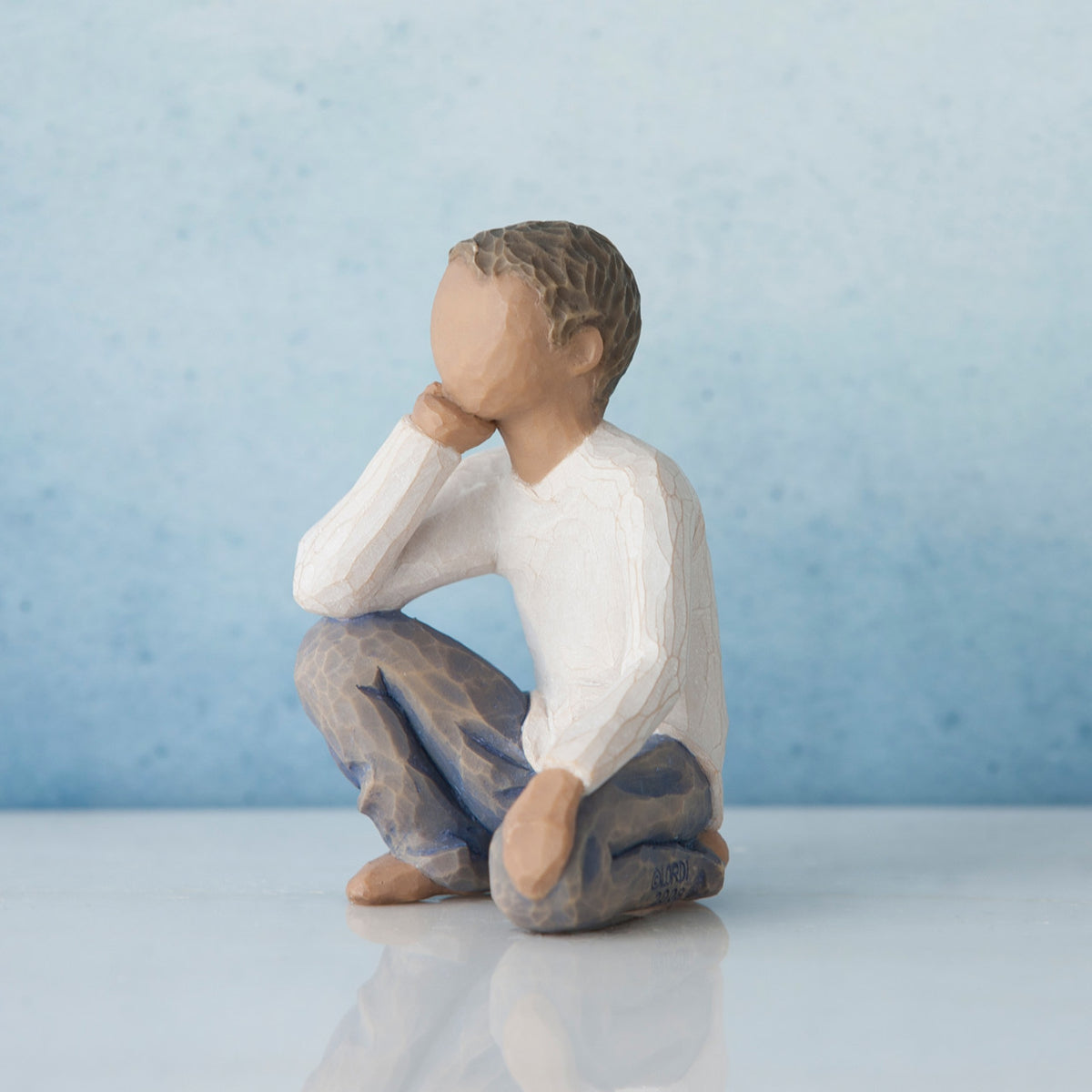 Small young boy figurine in white shirt and blue jeans with one hand up to his face kneeling - blue background