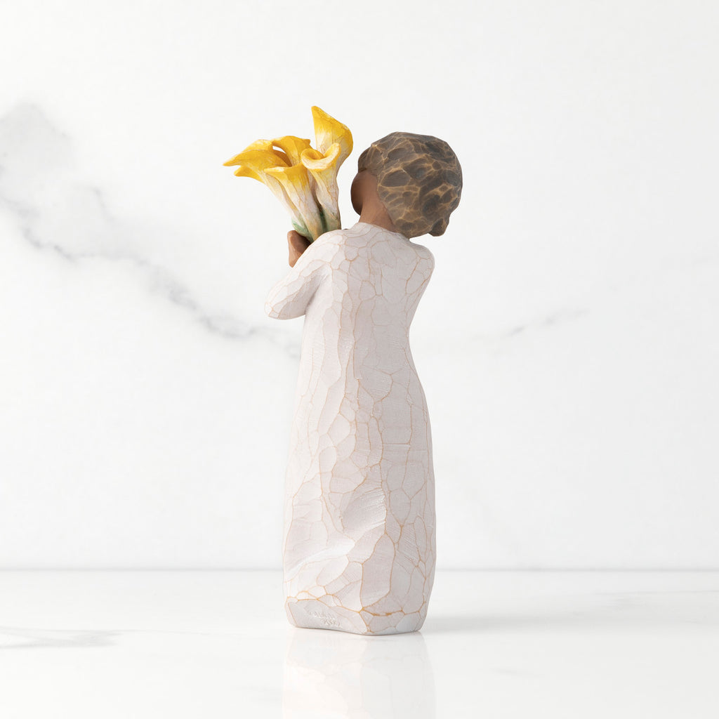 Back view of standing figure in cream dress with darker skin holding yellow calla lilies in her arms