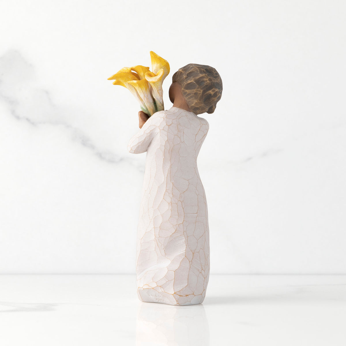 Back view of standing figure in cream dress with darker skin holding yellow calla lilies in her arms