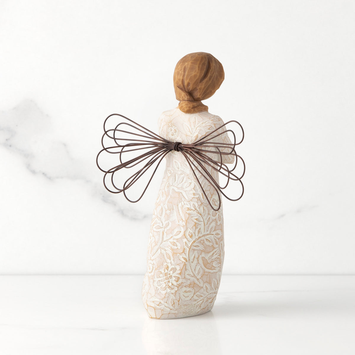 Back view of standing female figure with wire wings in cream dress carved with natural symbols, hands crossed under gold heart on chest