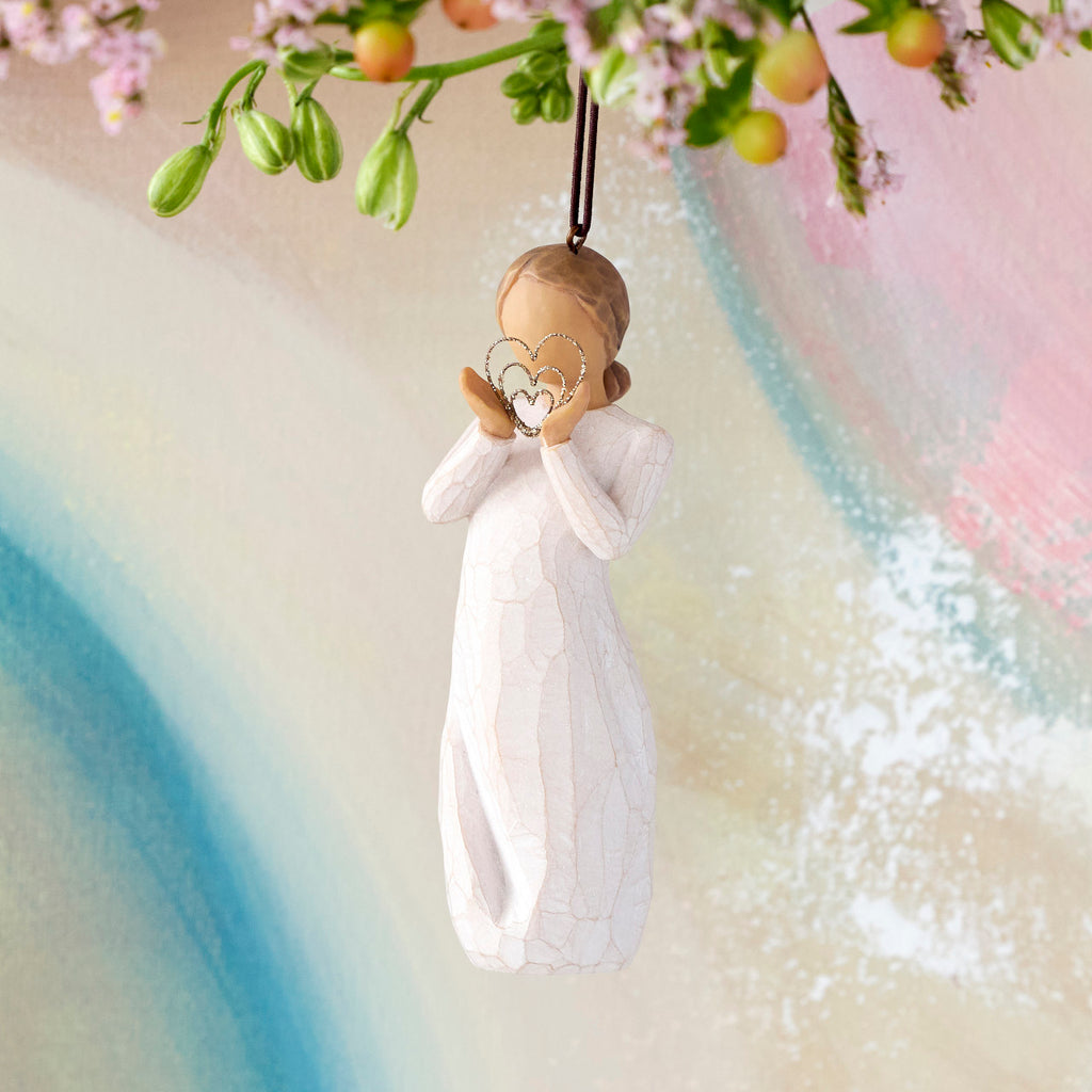 Willow Tree Lots of Love Ornament, 4.5”h hand-painted resin figure in cream dress, holding wire concentric heart with gold glitter, brown elastic cord, hanging in front of multi-pastel painting