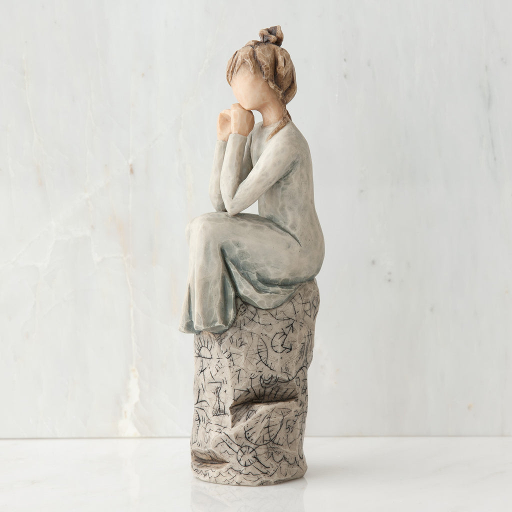 Brunette girl figurine in light grey dress sitting on grey rock facing the side - white background