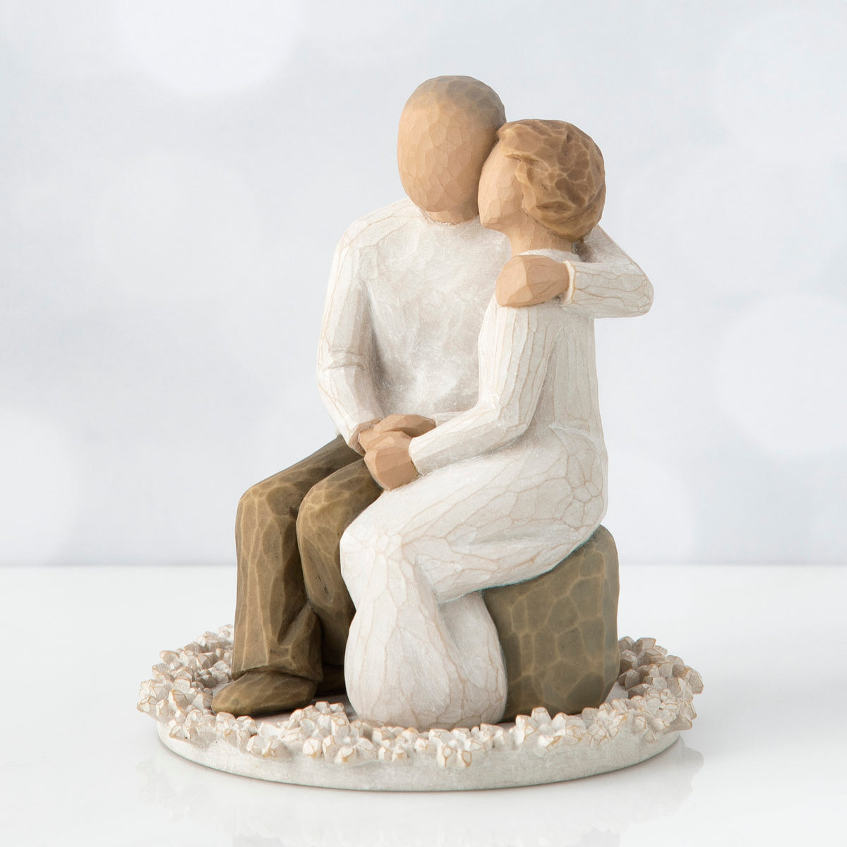 Man and woman figurine in white sitting on a rock on top of round white plaque hugging one another