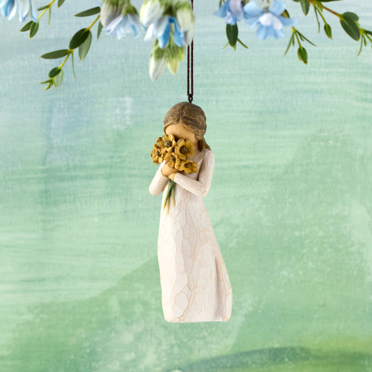 Willow Tree Warm Embrace Ornament, 4.25”h hand-painted resin figure in cream dress, holding bouquet of sunflowers up to face, brown elastic cord, hanging in front of blue green painting
