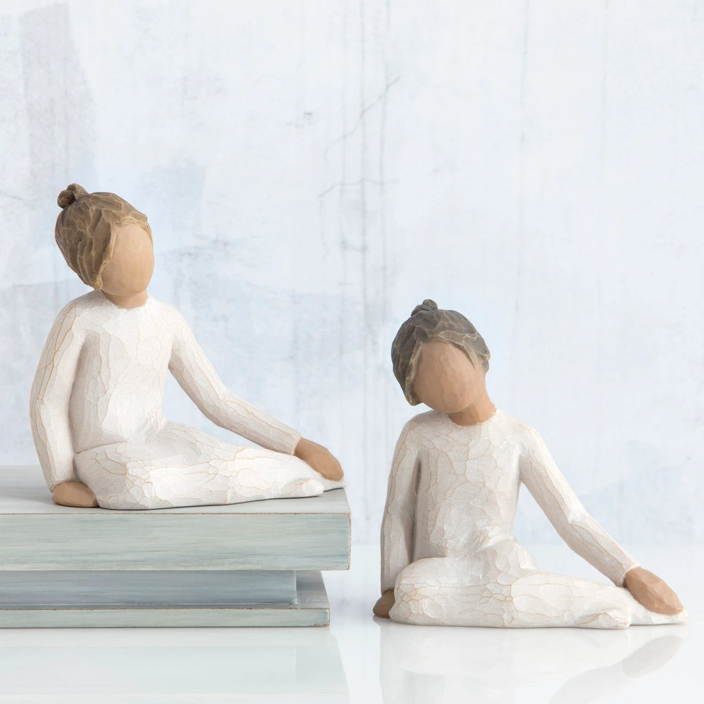 Two young brunette girl figurines in white sitting down next to wooden stack of books