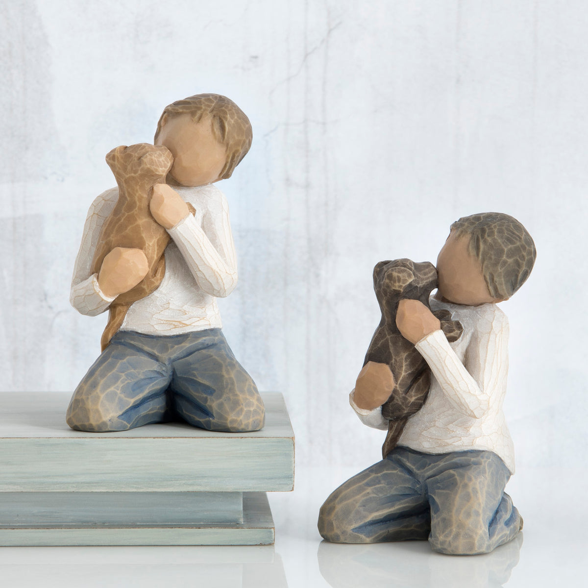Front view of boy with light brown hair wearing white shirt and jeans and holding a light brown puppy as it licks his face. Beside him, front view of boy with dark brown hair wearing white shirt and jeans and holding a dark brown puppy as it licks his face.