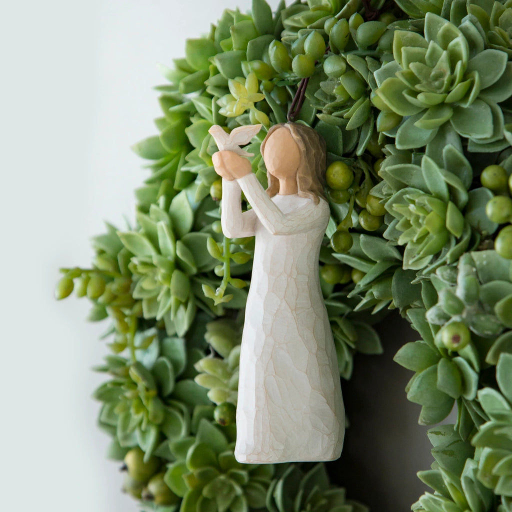 Willow Tree Soar Ornament, 4.5”h hand-painted resin figure in cream dress, holding white dove in extended arms, brown elastic cord, hanging in wreath of succulents
