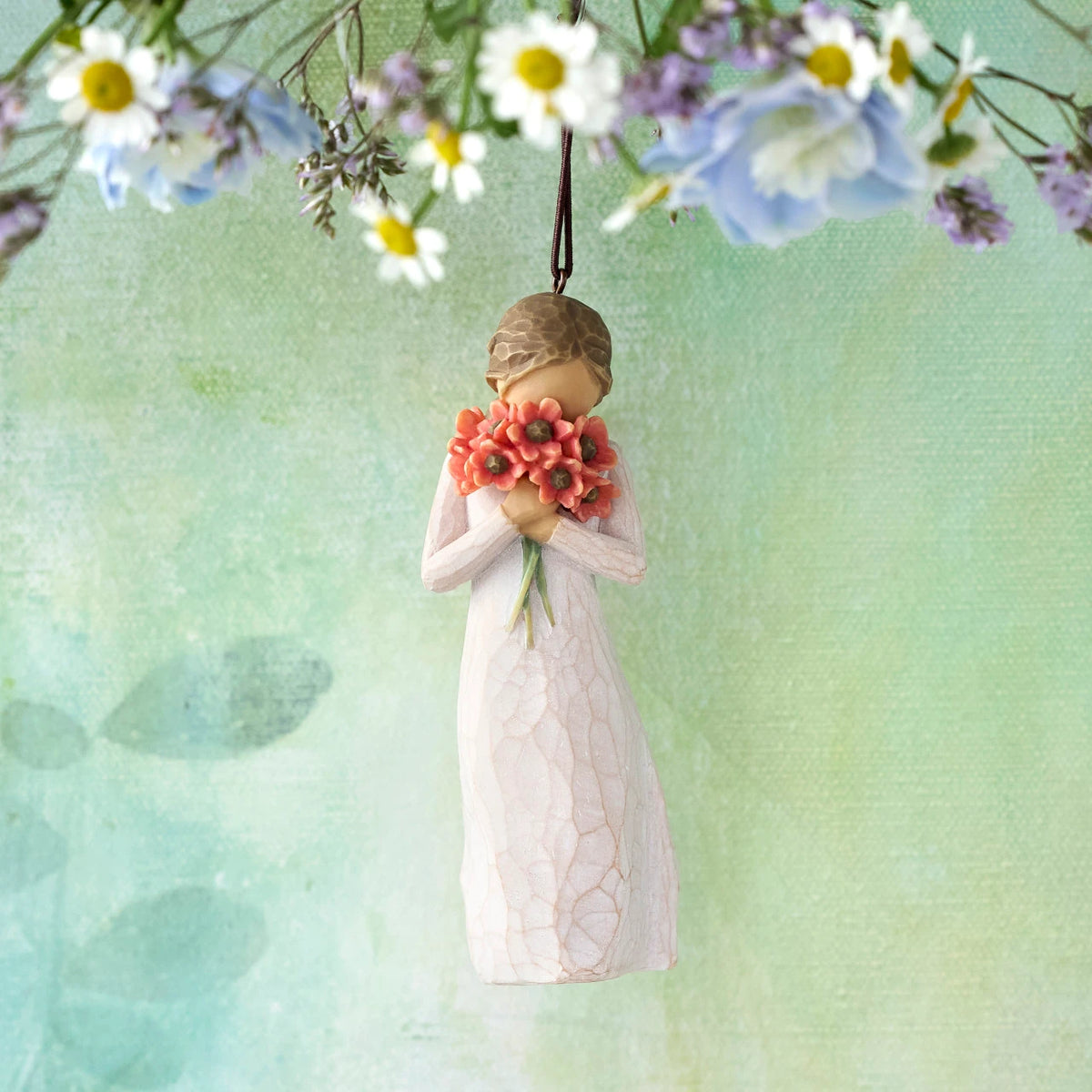 Willow Tree Surrounded by Love Ornament, 4”h hand-painted resin figure in cream dress holding bouquet of poppies to face, brown elastic cord, hanging in front of pale green painting