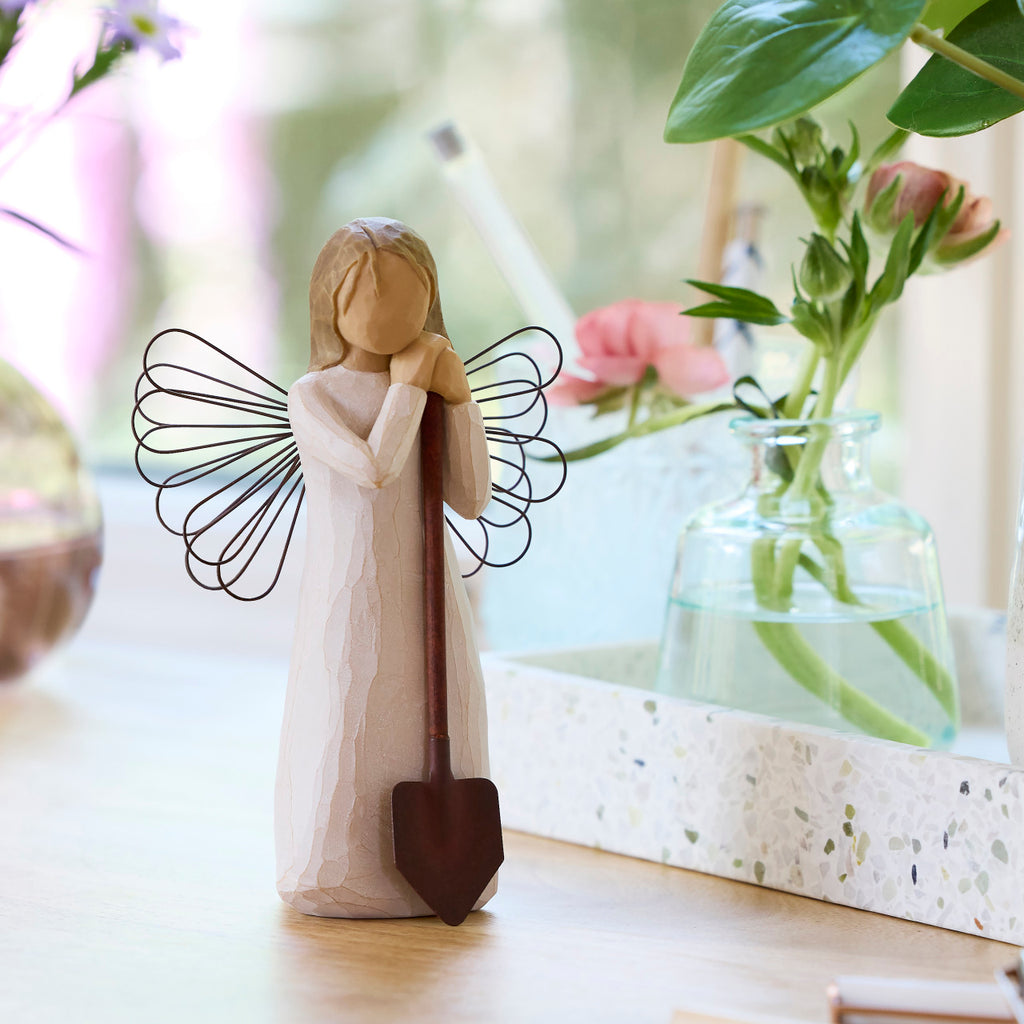 Willow Tree Angel of the Garden, 5.5”h hand-painted standing resin figure in cream dress with wire wings, leaning against brown metal shovel, on table with florals