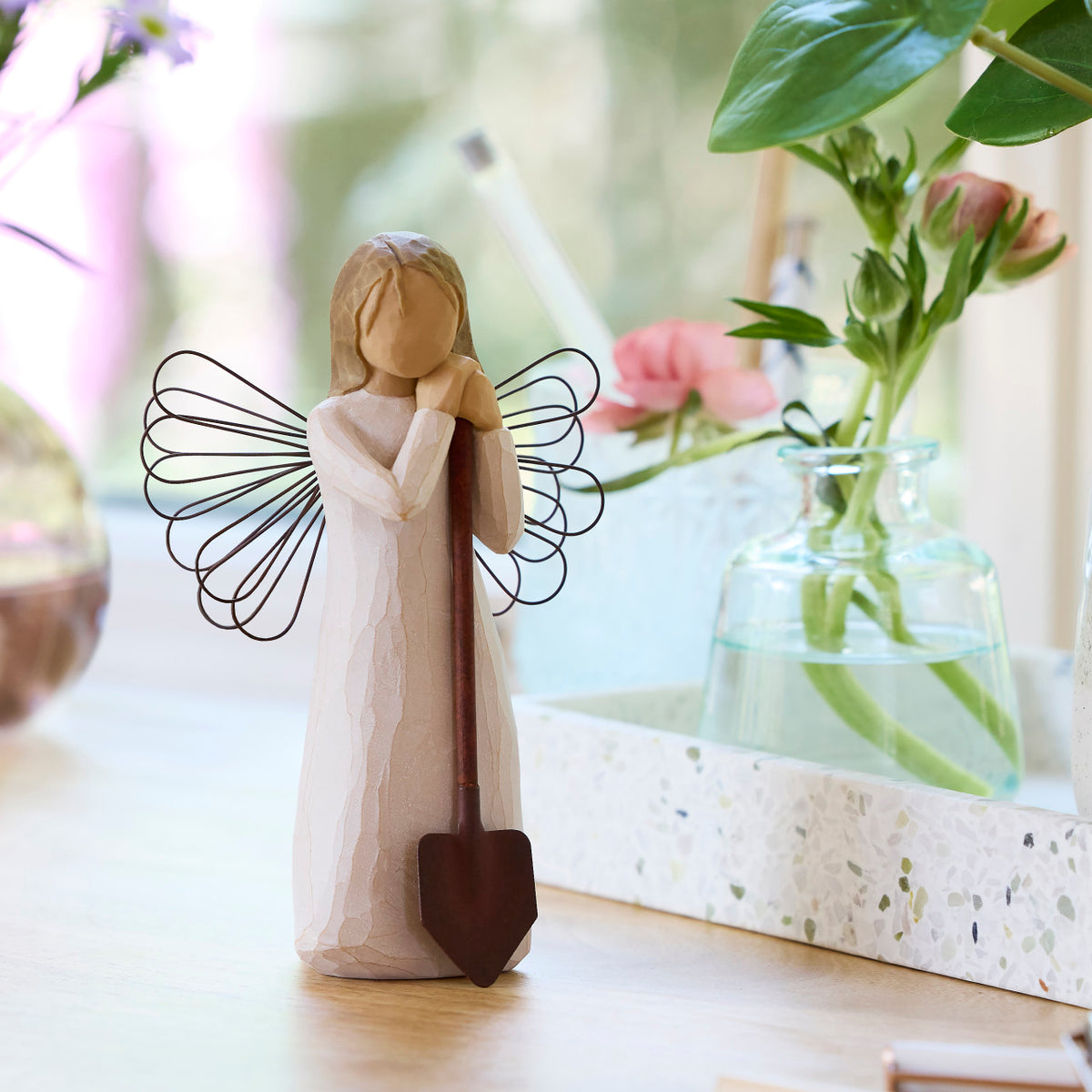 Willow Tree Angel of the Garden, 5.5”h hand-painted standing resin figure in cream dress with wire wings, leaning against brown metal shovel, on table with florals