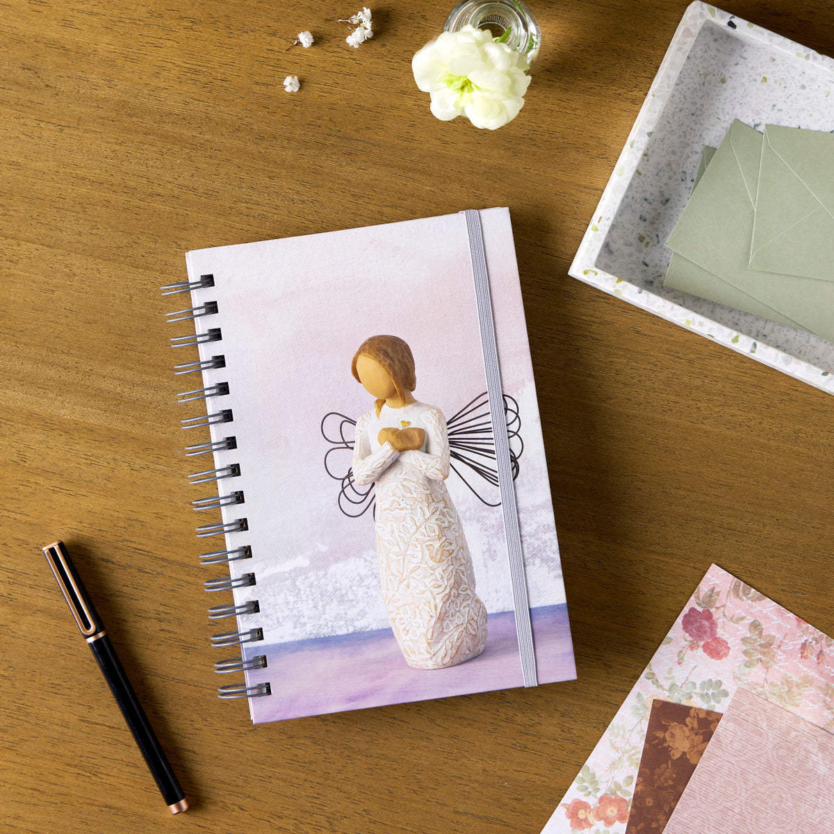 Willow Tree Remembrance Notebook, 6.5"w x 8.5"h, hard-cover, spiral-bound, 240 lined pages, front cover features Remembrance figure on background of purple-blue painting, closed on desk