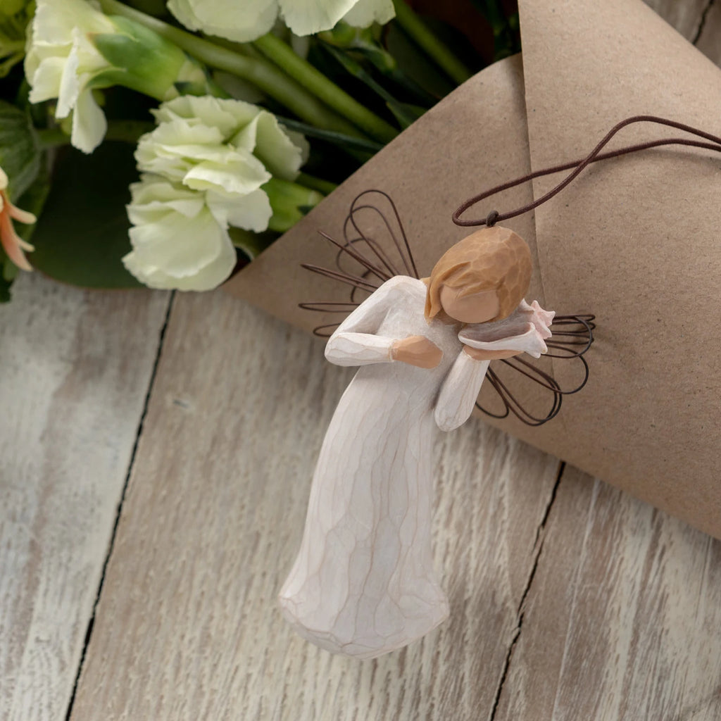 Willow Tree Thinking of You Ornament, 4.5”h hand-painted resin angel figure in cream dress with wire wings, holding pink conch shell to ear, brown elastic cord, laying next to floral bouquet