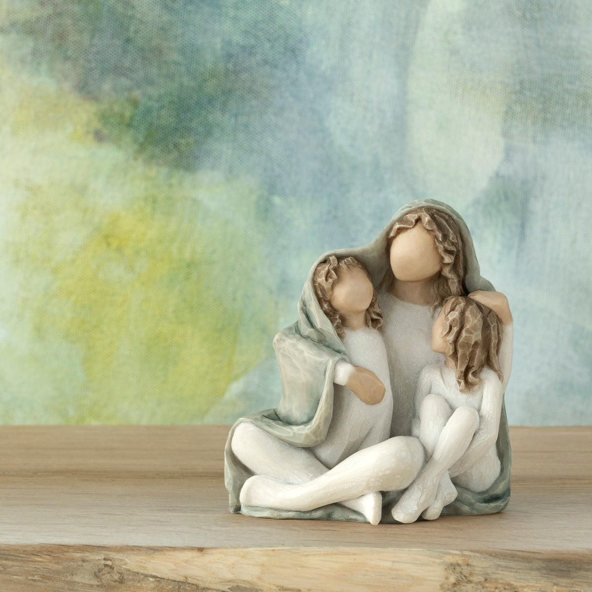 Willow Tree Cozy, 3.5”h hand-painted resin female figure sitting with arms around two children, all in cream clothes and wrapped in blue-gray blanket, in front of blue painting