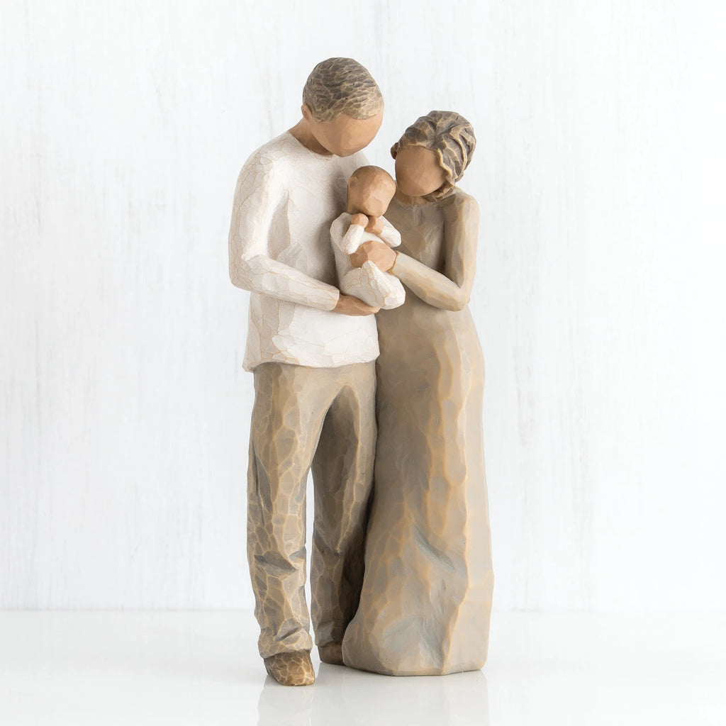 Man and woman faceless figurines holding baby in white outfit - man is dressed in white shirt and brown pants, woman is dressed in brown dress