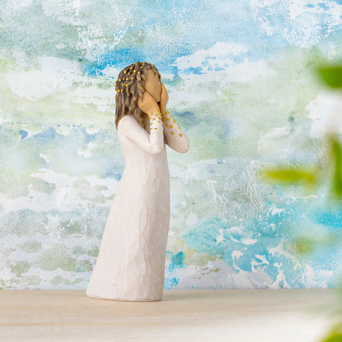 Willow Tree Littlest Christmas Angel, 5.5”h hand-painted standing resin figure in cream dress, both hands up to face in awe, gold leaf dots on sleeves and hair, in front of blue-white painting