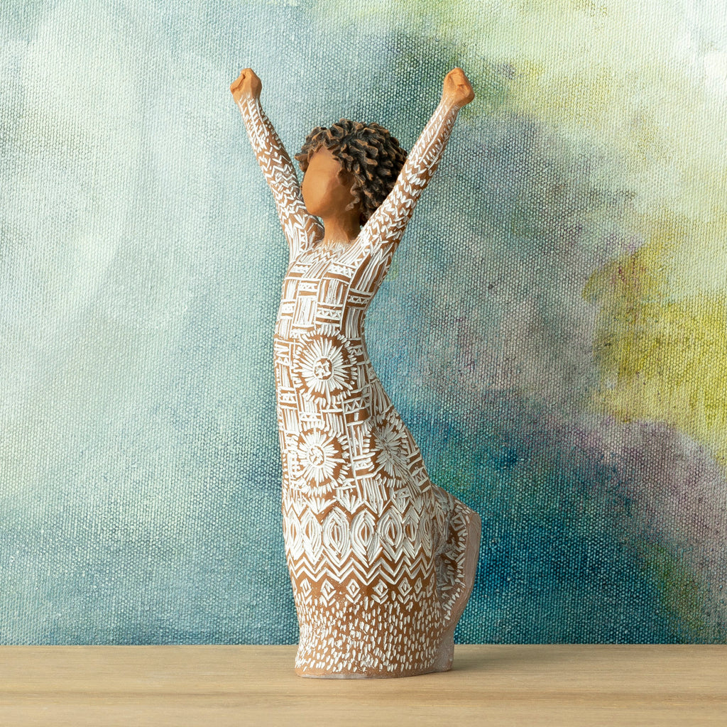 Willow Tree Courageous Joy, 7”h hand-painted resin figure with darker skin and natural hairstyle, standing, both arms raised overhead, cream dress etched with textile patterns, in front of deep teal painting