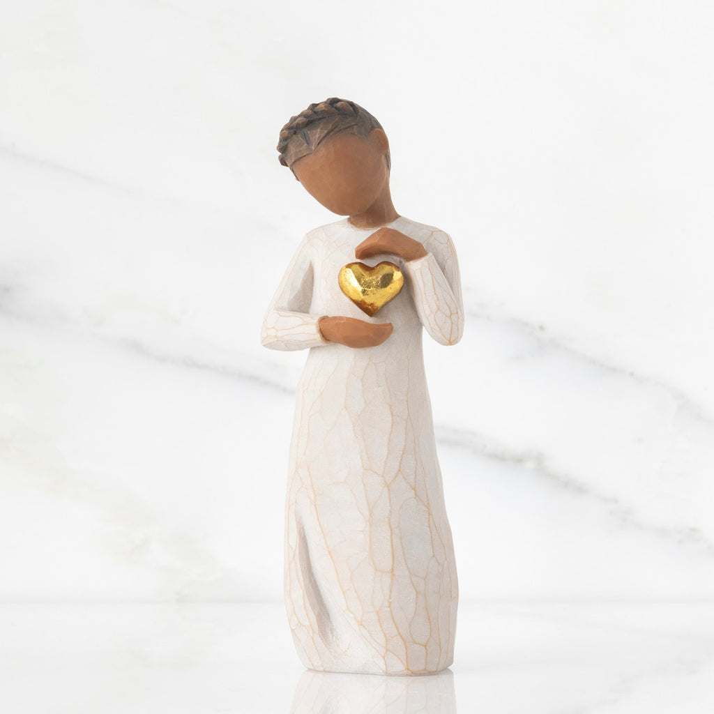 Front view: standing figure in cream dress with darker skin and dark hair braid, framing gold leaf heart with both hands on chest