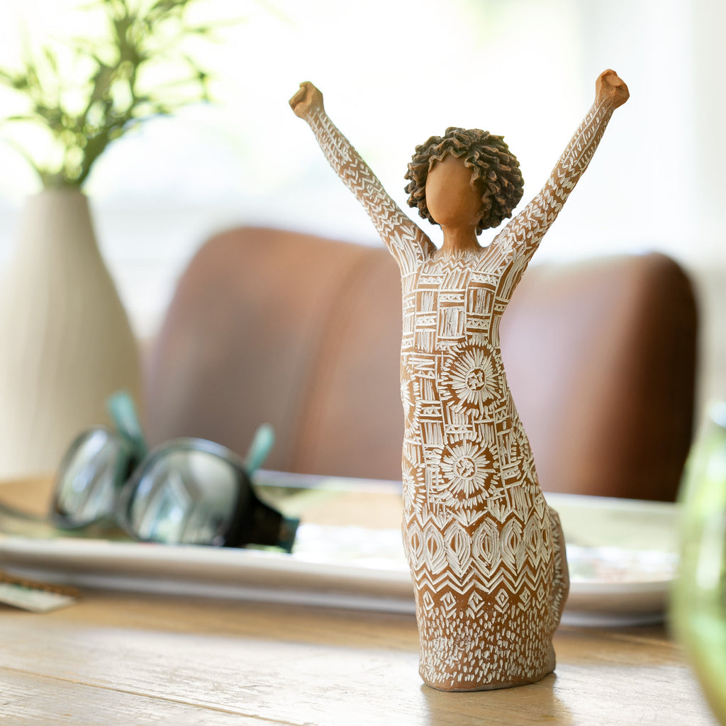 Willow Tree Courageous Joy, 7”h hand-painted resin figure with darker skin and natural hairstyle, standing, both arms raised overhead, cream dress etched with textile patterns, on coffee table
