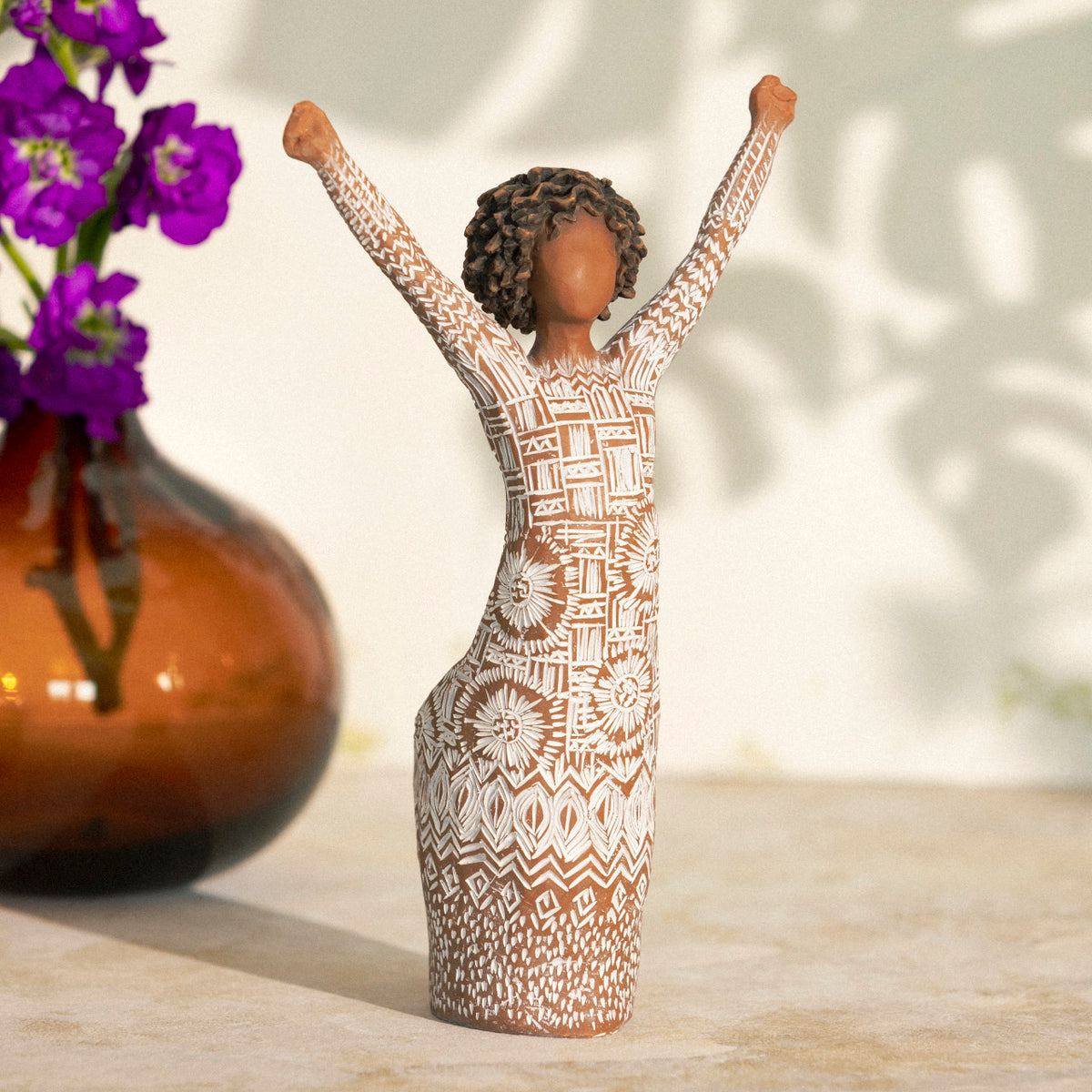 Willow Tree Courageous Joy, 7”h hand-painted resin figure with darker skin and natural hairstyle, standing, both arms raised overhead, cream dress etched with textile patterns, on table in bright light