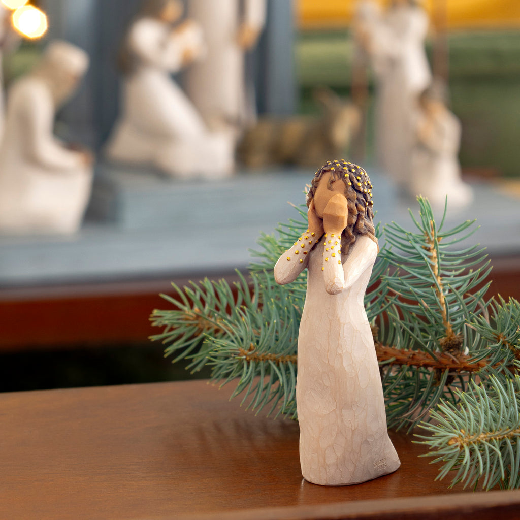Willow Tree Littlest Christmas Angel, 5.5”h hand-painted standing resin figure in cream dress, both hands up to face in awe, gold leaf dots on sleeves and hair, on table with Nativity